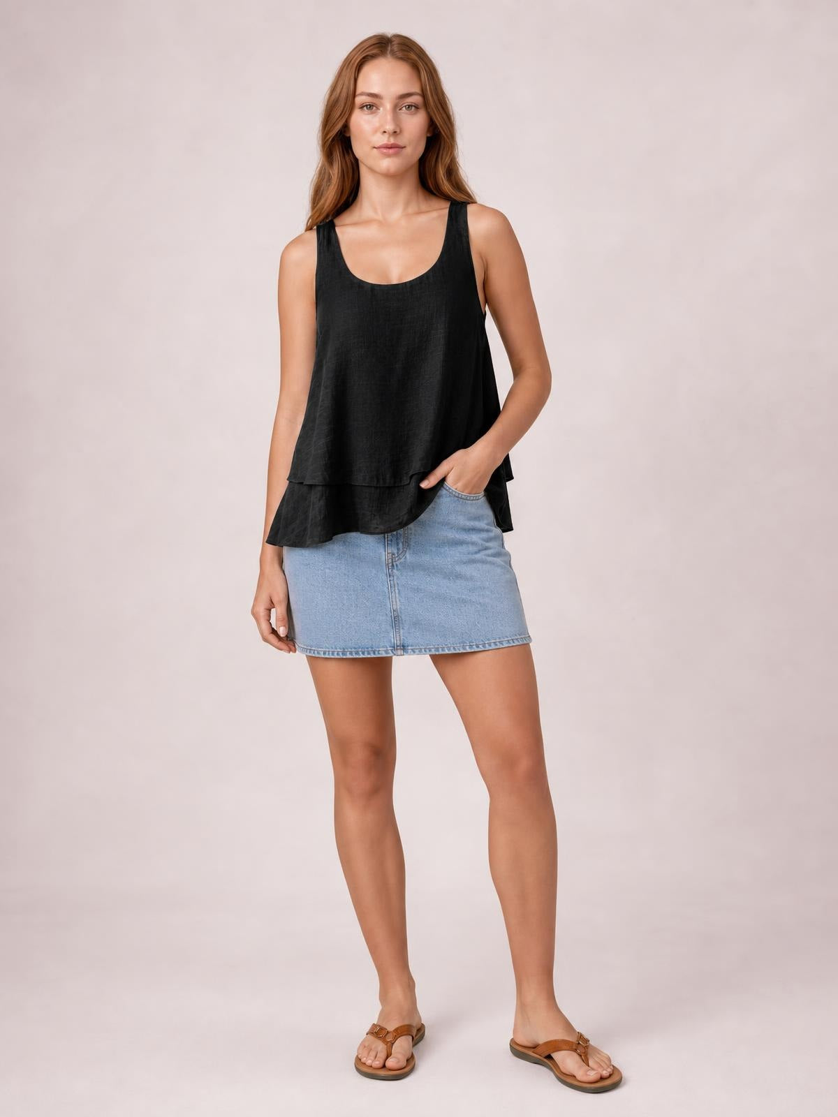Model poses in a black sheer wide-strap layered tank with a loose tiered silhouette and curved hem, paired with a light blue denim mini skirt.
