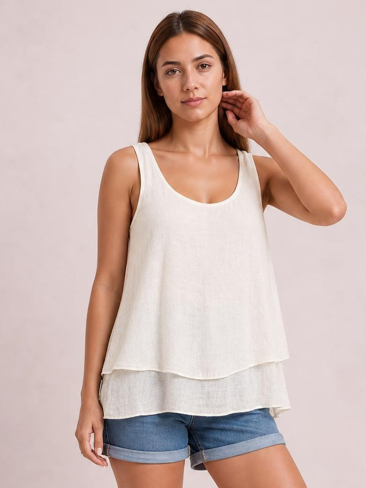A model wears an ivory sheer layered tank with wide straps, a scoop neckline, and a relaxed flowing silhouette, touching her hair with one hand.