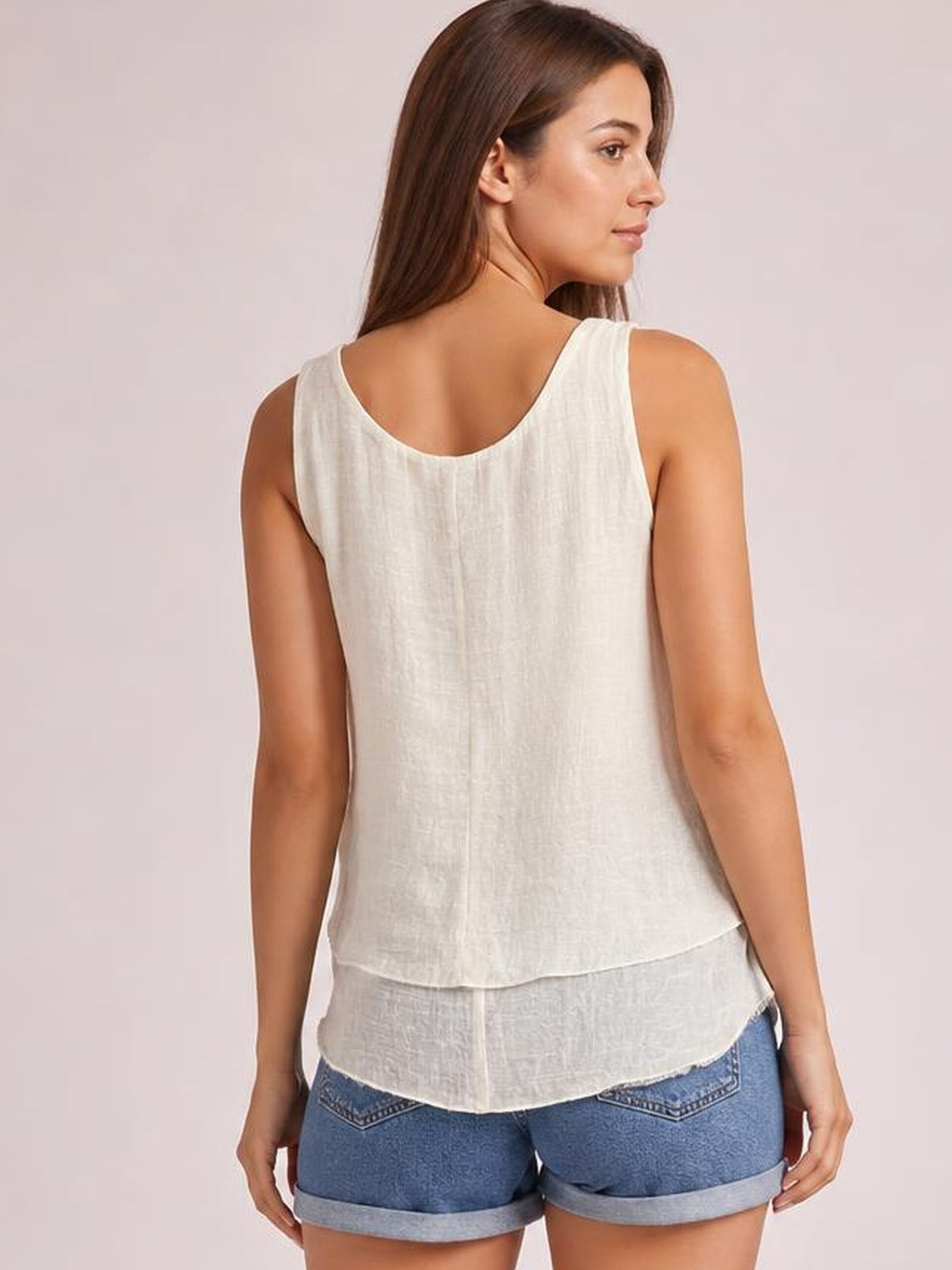 Model shown from the back wearing a white wide-strap layered tank with a sheer, relaxed silhouette and curved hem over denim shorts.