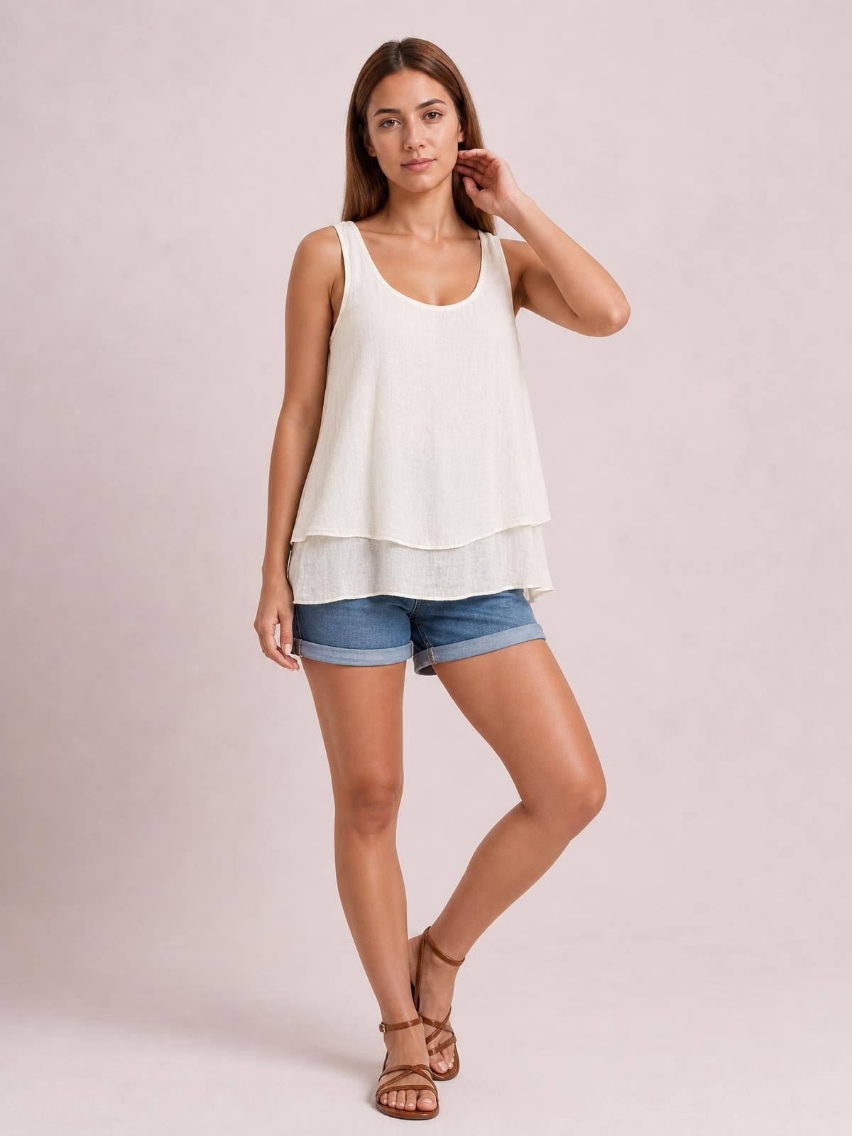 Model poses with one hand near her hair, wearing a white sheer wide-strap layered tank with a scoop neckline, loose silhouette, and tiered curved hems.