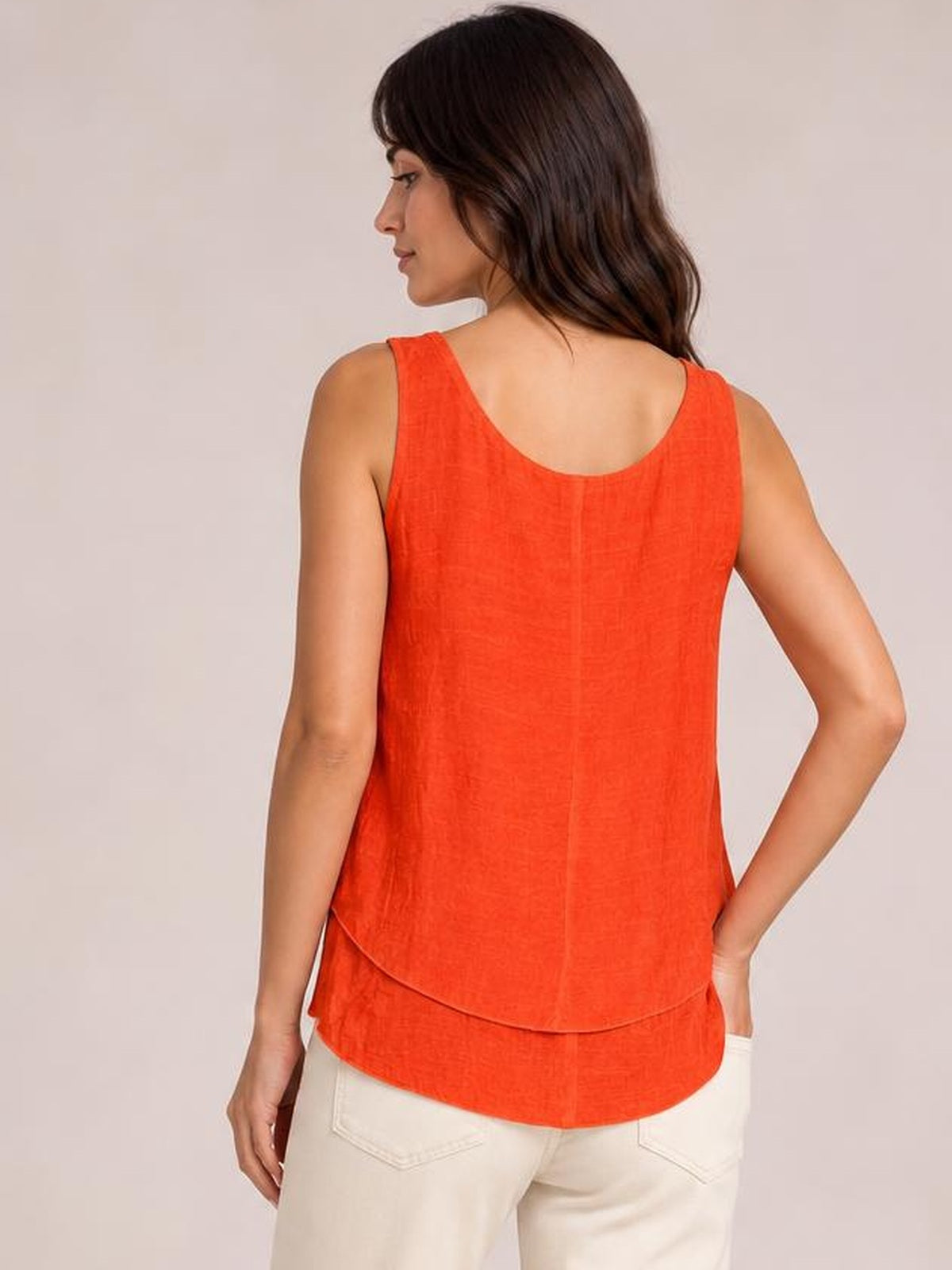 A model poses with one hand on her hip, showing the back of an orange wide-strap layered tank with a relaxed silhouette and curved double hem.