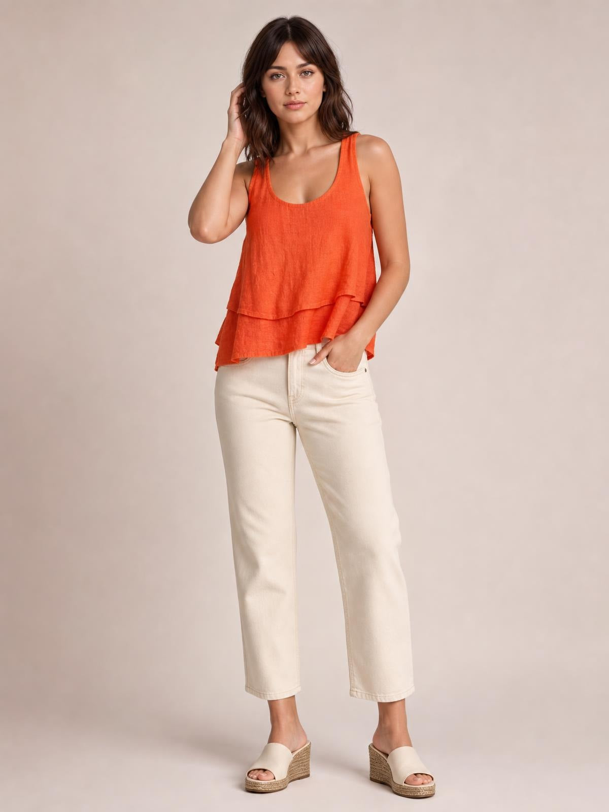 A model poses in an orange wide-strap layered tank with a sheer, relaxed silhouette and two flowing tiers, paired with cropped cream pants.