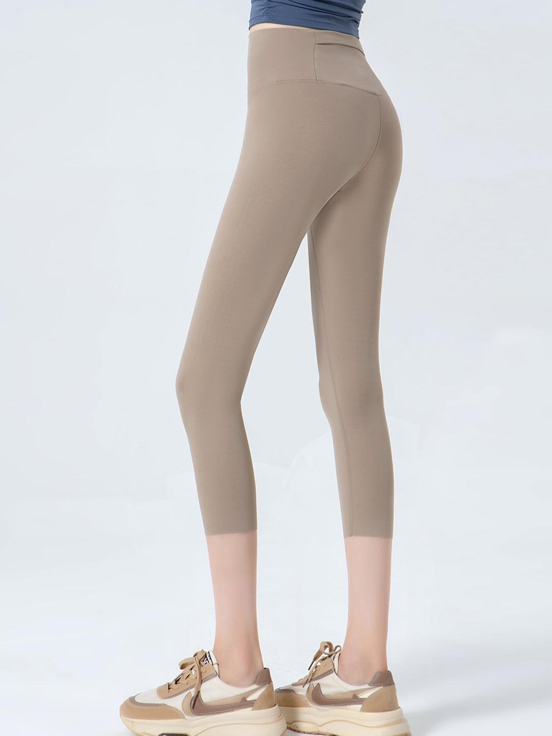 A model stands with legs slightly apart in taupe cropped sports leggings featuring a wide waistband, opaque stretch fabric, and smooth contoured seams.