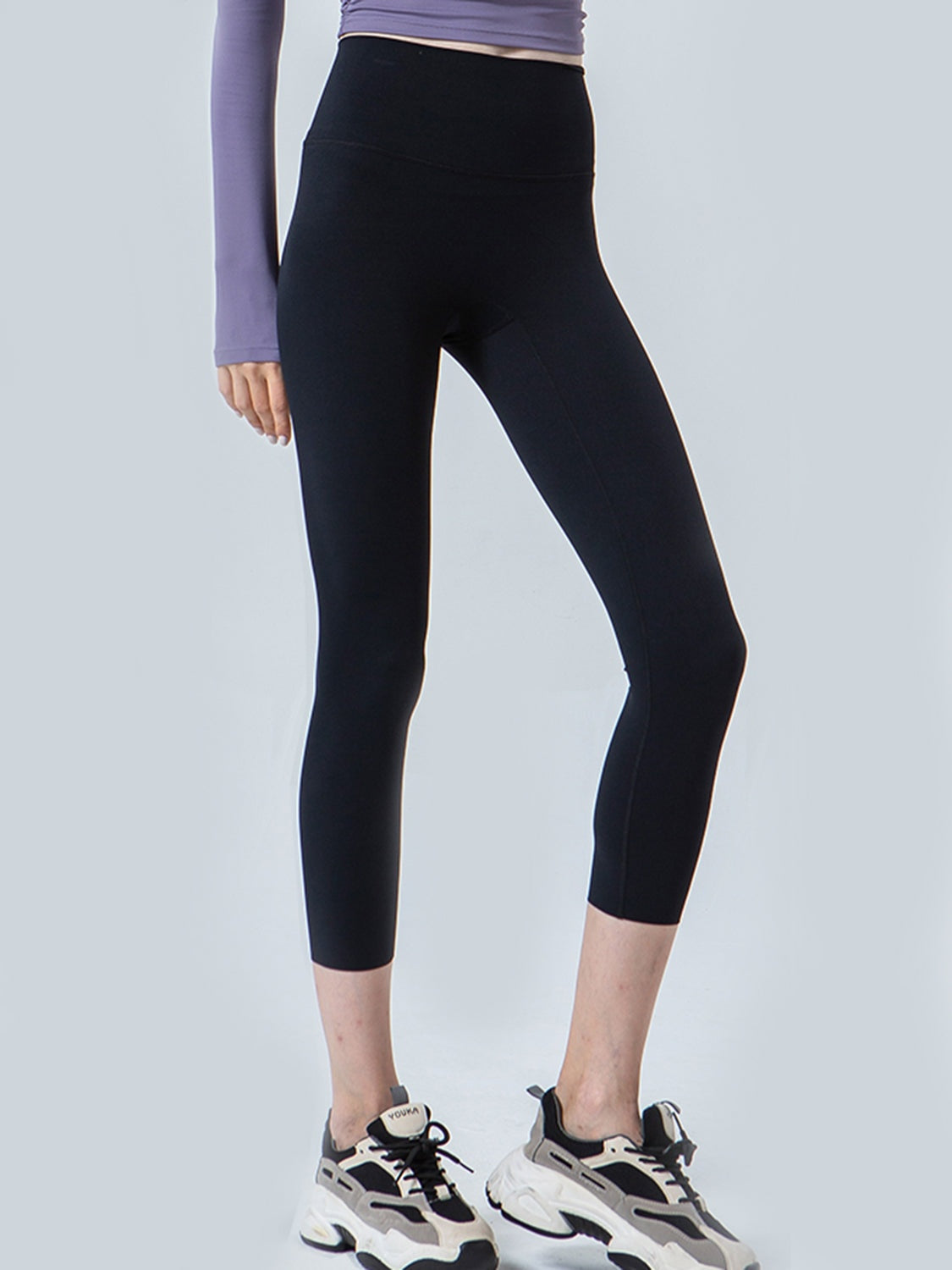Model wears black cropped sports leggings with a wide waistband and fitted silhouette, standing with one leg angled forward.
