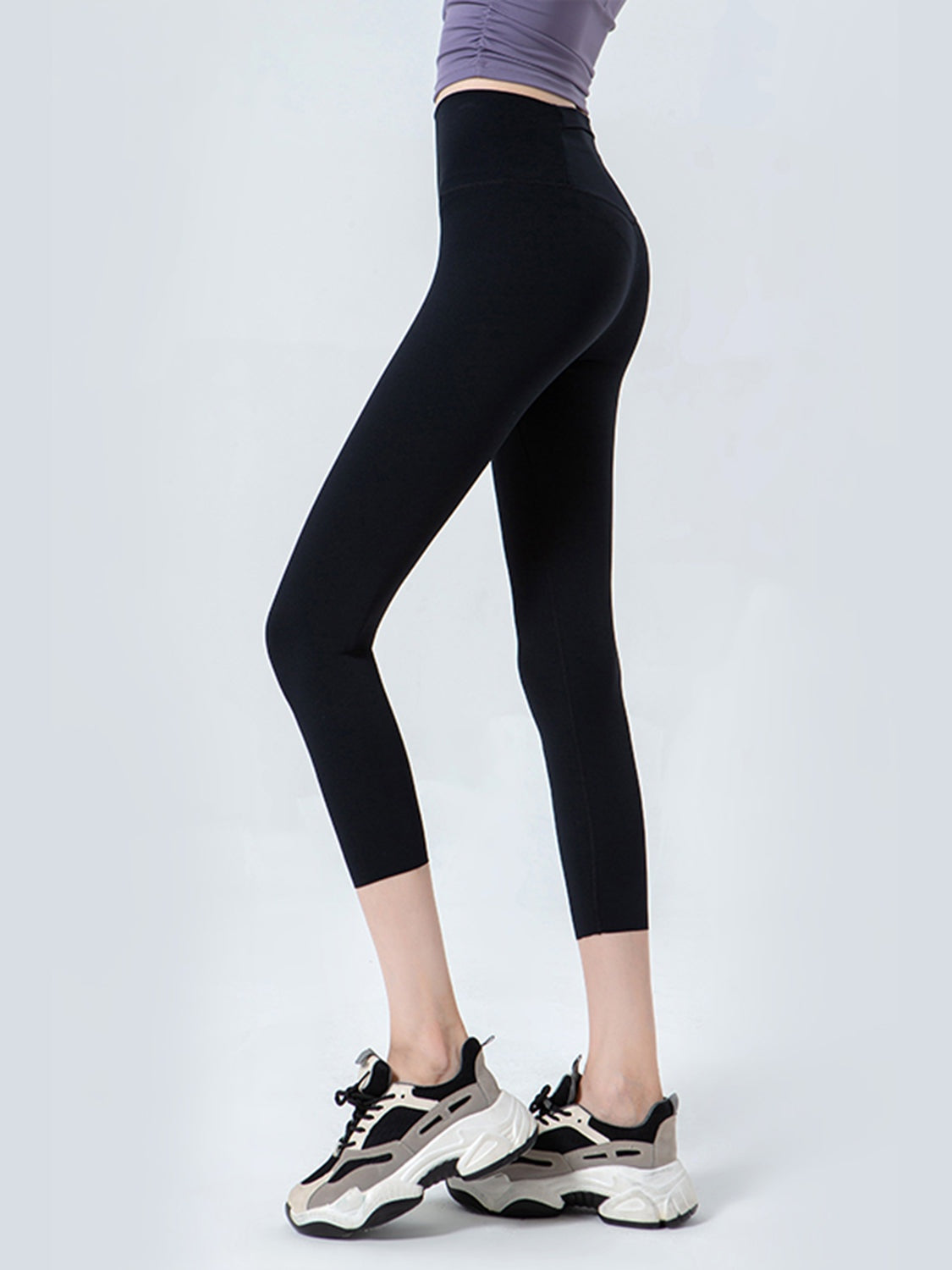 A model poses in profile wearing black cropped sports leggings with a wide waistband and sleek, close-fitting stretch silhouette ending below the knees.