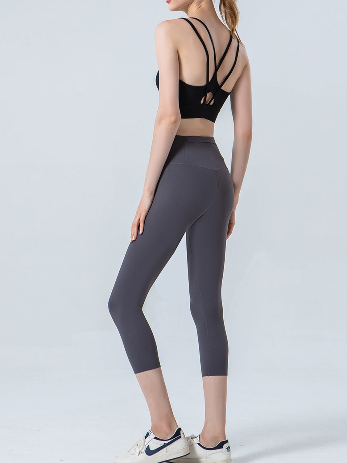 Model in a rear three-quarter pose wears dark gray cropped sports leggings with a wide waistband, fitted silhouette, and curved seam construction.