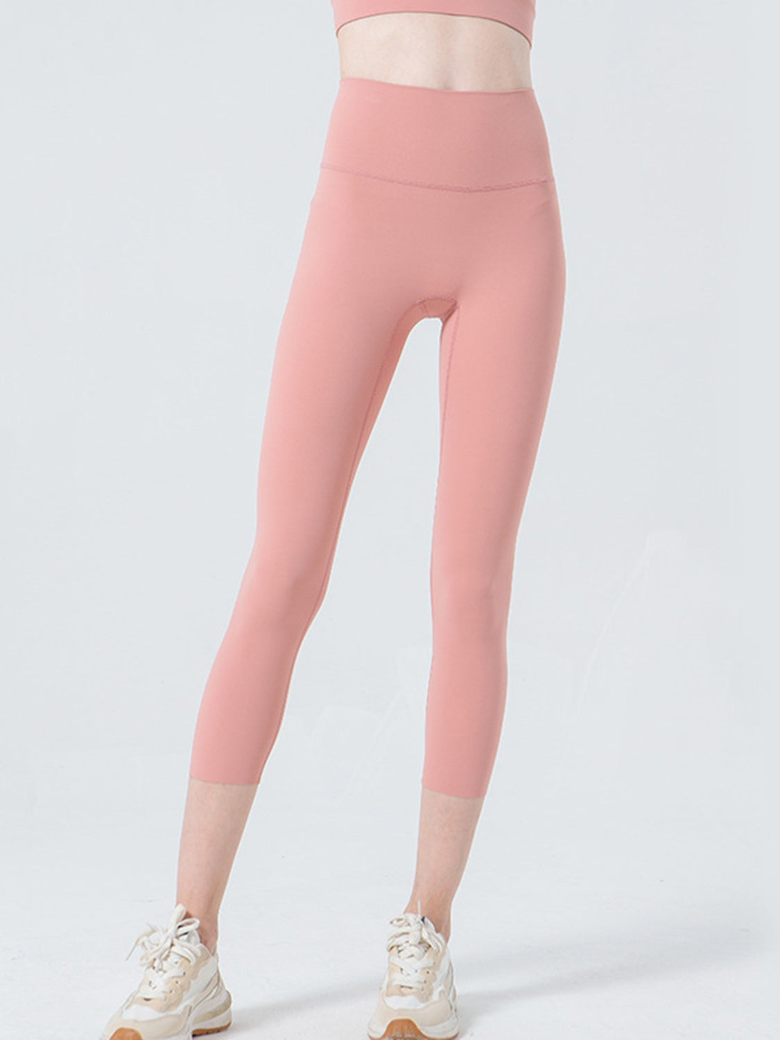 A model stands with one leg slightly bent in pink cropped sports leggings featuring a wide waistband, fitted silhouette, and visible front seam construction.
