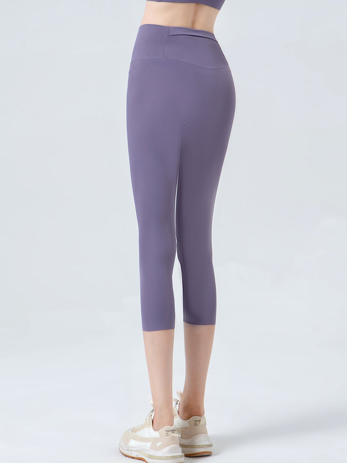 A model stands with one leg crossed behind the other, wearing purple cropped sports leggings with a wide waistband, fitted silhouette, and curved seam detailing.