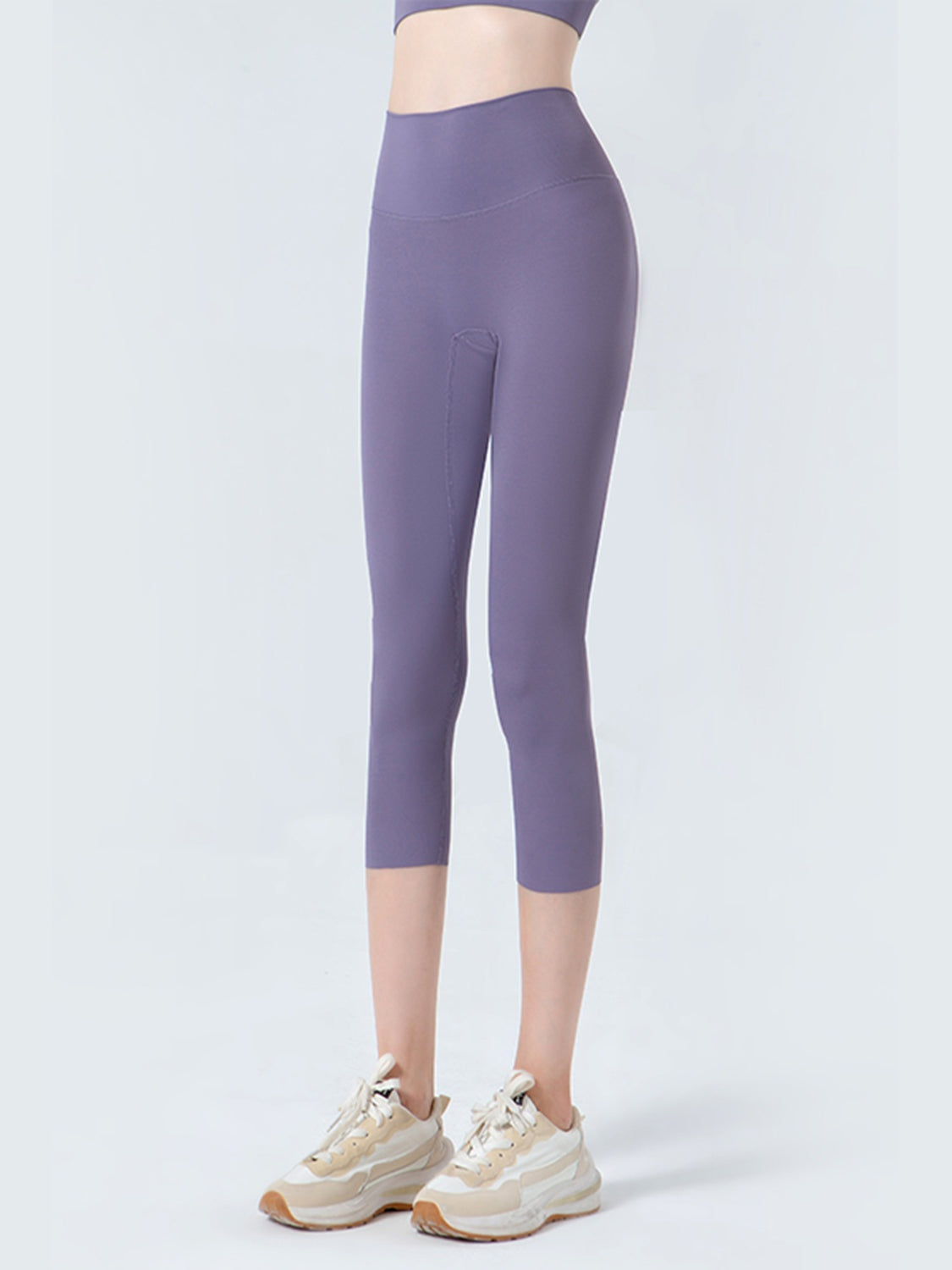 Model stands in purple cropped sports leggings with a wide waistband, fitted silhouette, opaque stretch fabric, and visible curved seam construction.