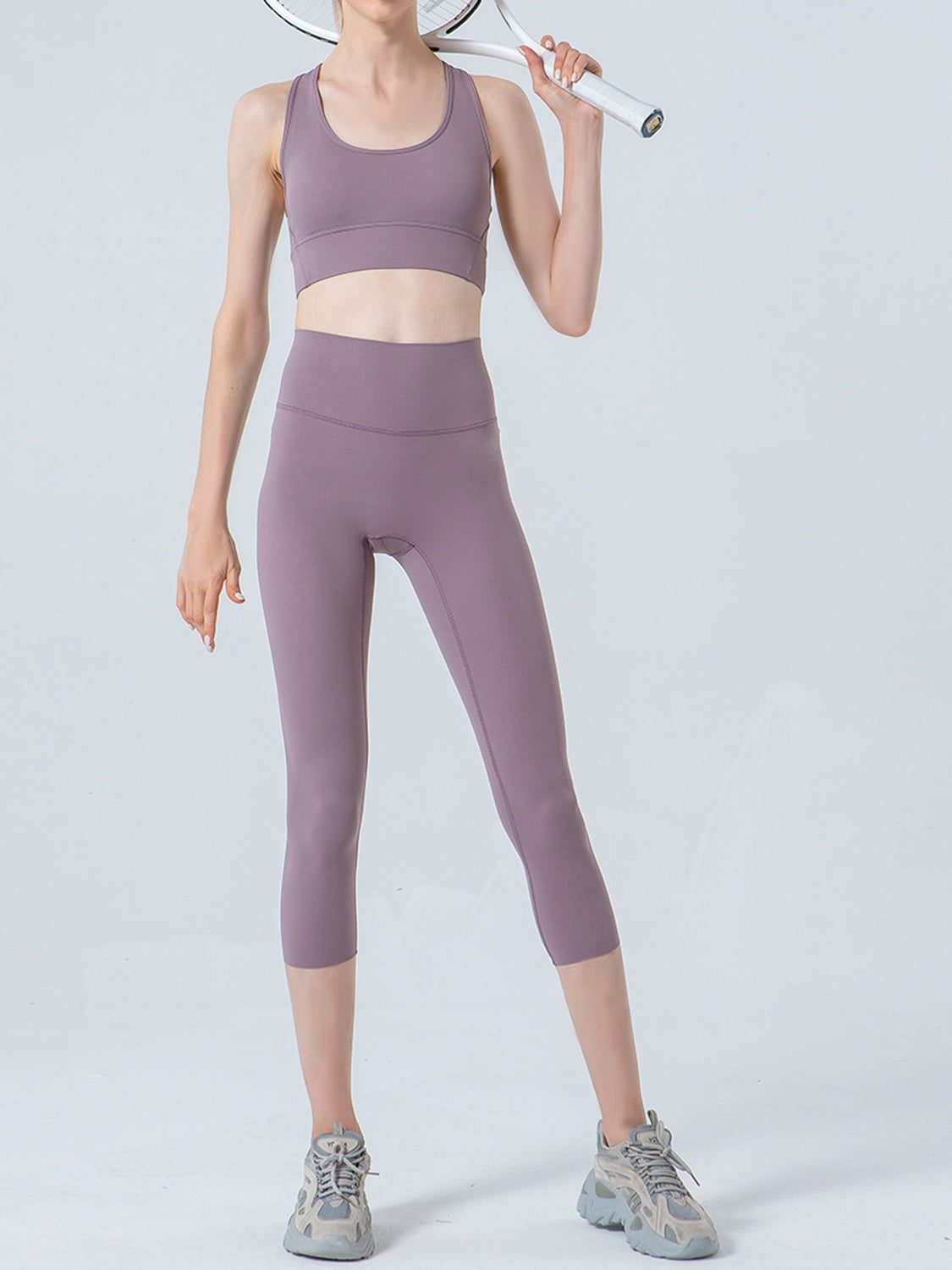Model wearing mauve wide-waistband cropped sports leggings with a fitted, seamed silhouette ending mid-calf, standing with one hand holding a tennis racket.
