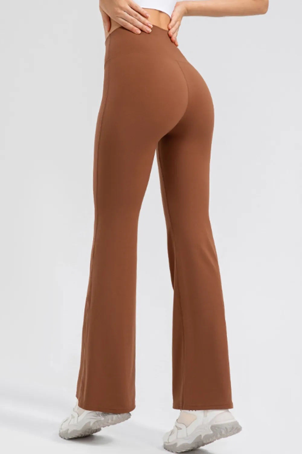 Model shown from behind adjusts the wide waistband of brown high-waist active leggings, featuring a smooth, fitted silhouette that flares into bootcut hems.