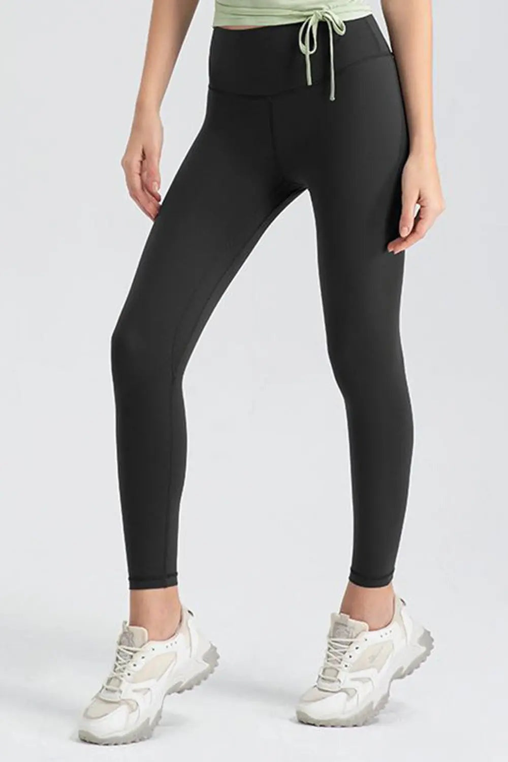 A cropped model stands with legs apart in black slim-fit active leggings featuring a wide waistband, smooth stretch fabric, and ankle-length tapered legs.