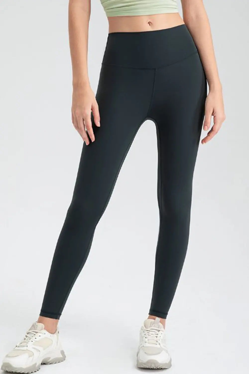 Model wears black wide-waistband active leggings in a slim, stretchy ankle-length silhouette with a smooth front and center seam, standing with legs apart.