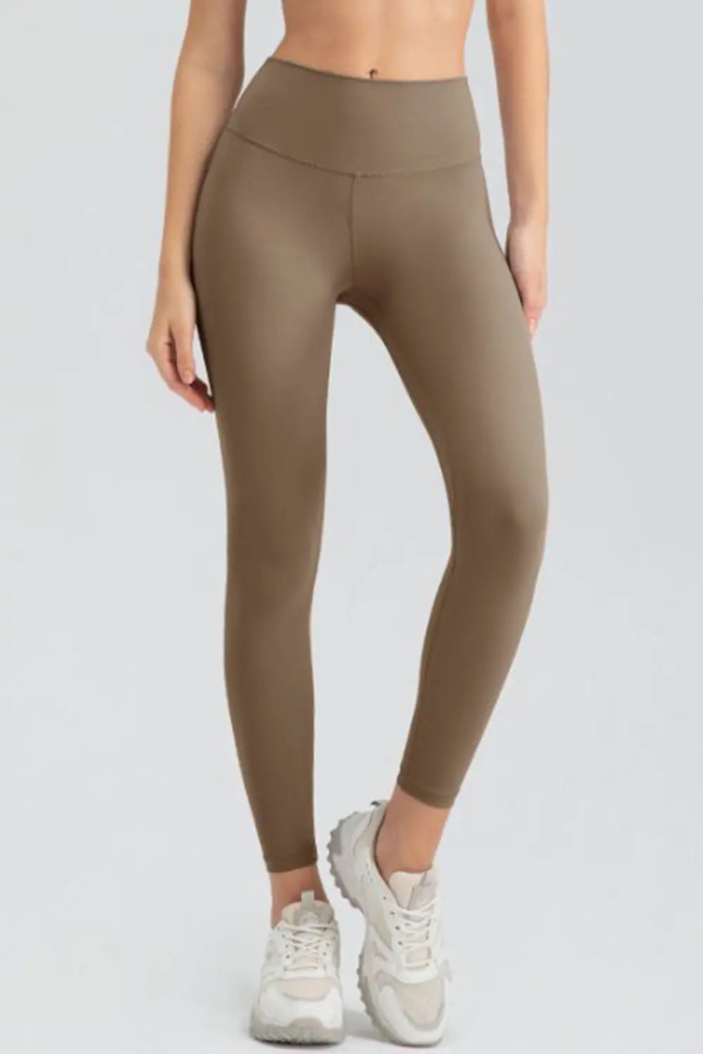 Model wears brown high-waisted slim-fit active leggings with a wide waistband, ankle-length silhouette, and visible front seam.