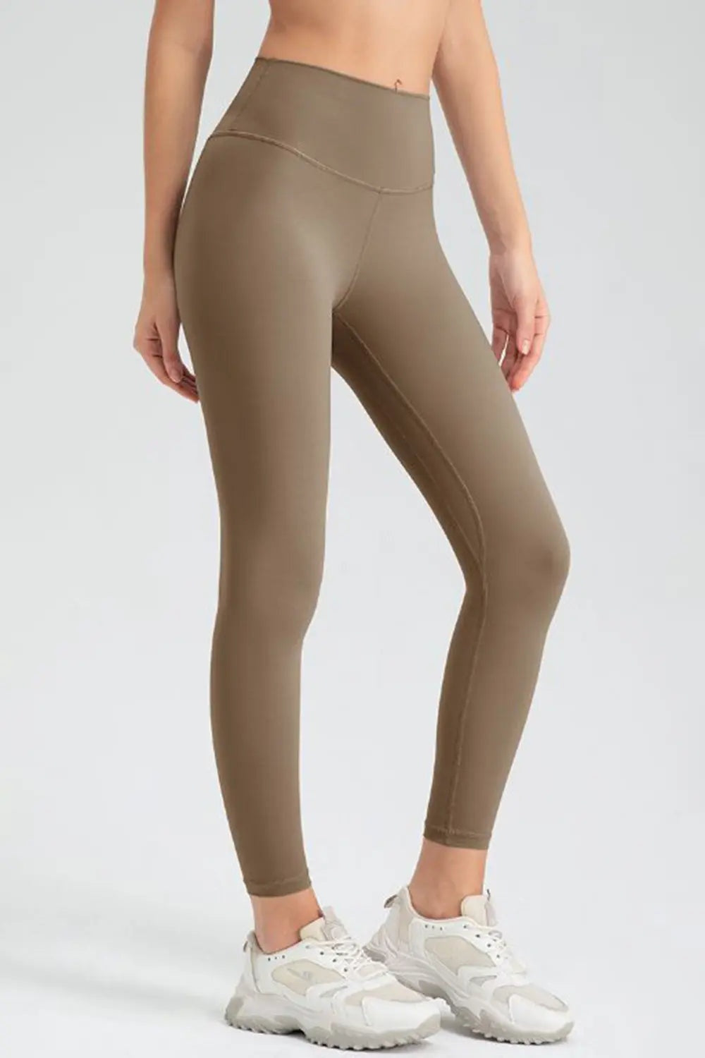 Model wears taupe slim-fit active leggings with a wide waistband, ankle-length silhouette, and visible curved and vertical seam construction while standing with one leg bent.