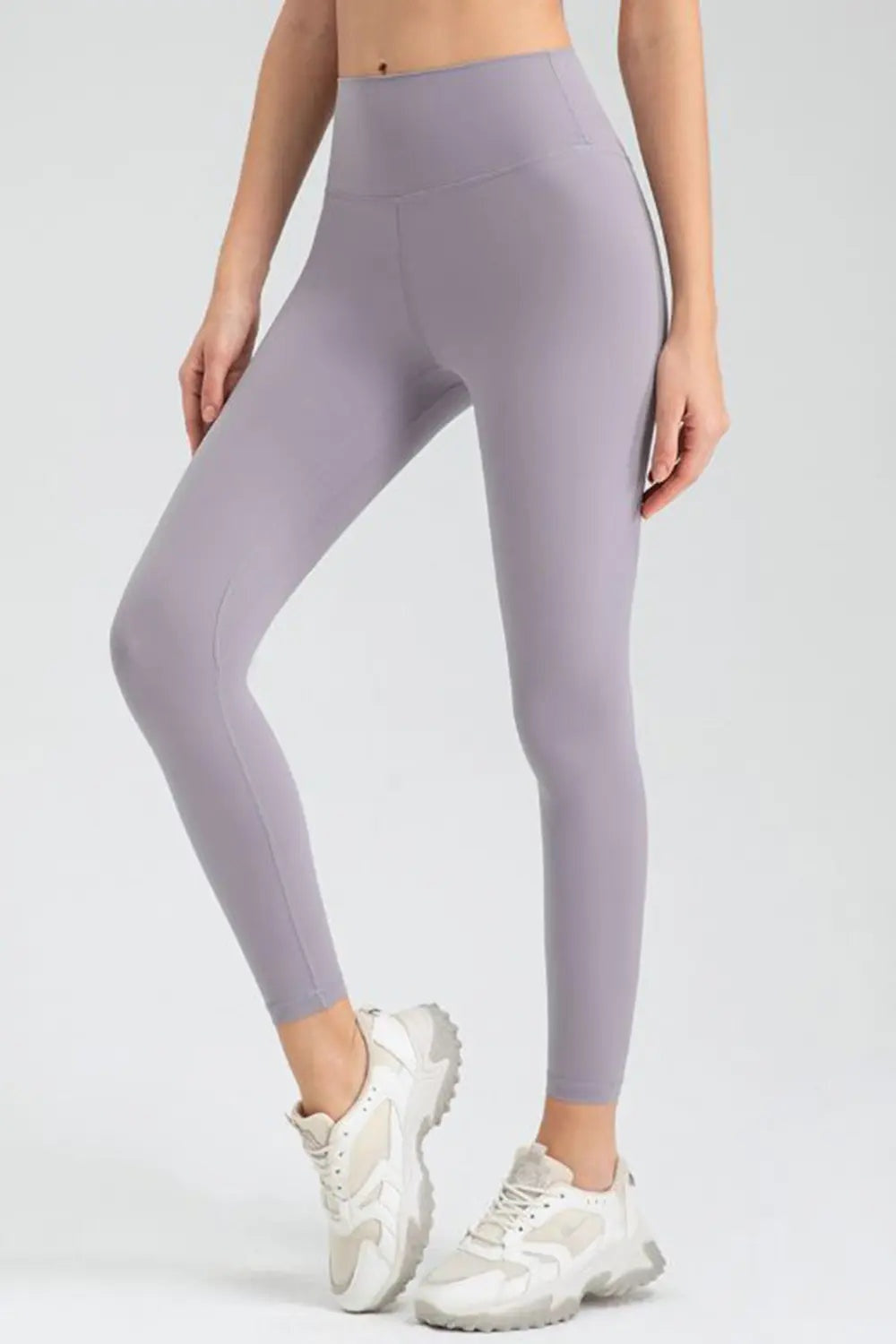 Model poses in lavender wide-waistband slim-fit active leggings with a high-rise silhouette and ankle-length construction, one knee bent.