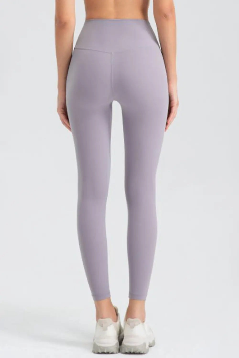 Rear view of a model standing with arms at her sides in lavender wide-waistband slim-fit active leggings, featuring a high-rise silhouette and full-length tapered legs.