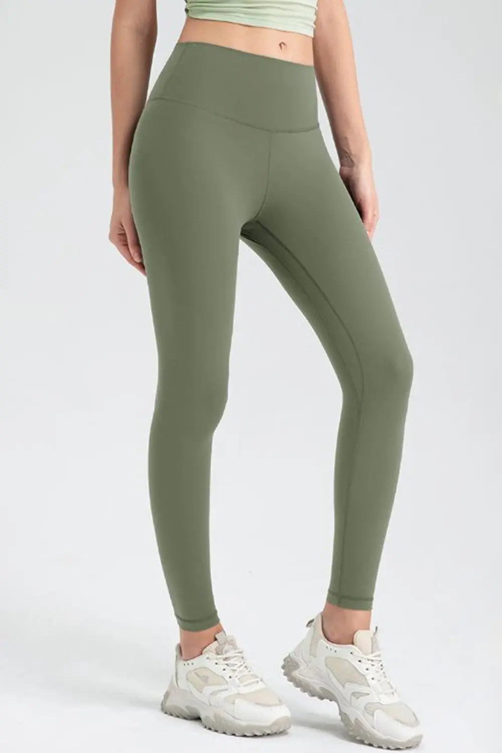 Model wears olive green wide-waistband slim-fit active leggings with a high-rise silhouette and visible seam construction, standing with one leg angled forward.