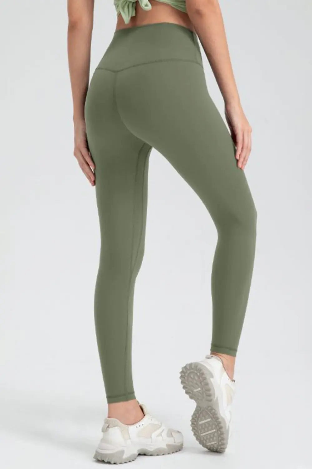 Model in a three-quarter rear pose wears olive green slim-fit active leggings with a wide waistband, full-length silhouette, and visible curved seam construction.