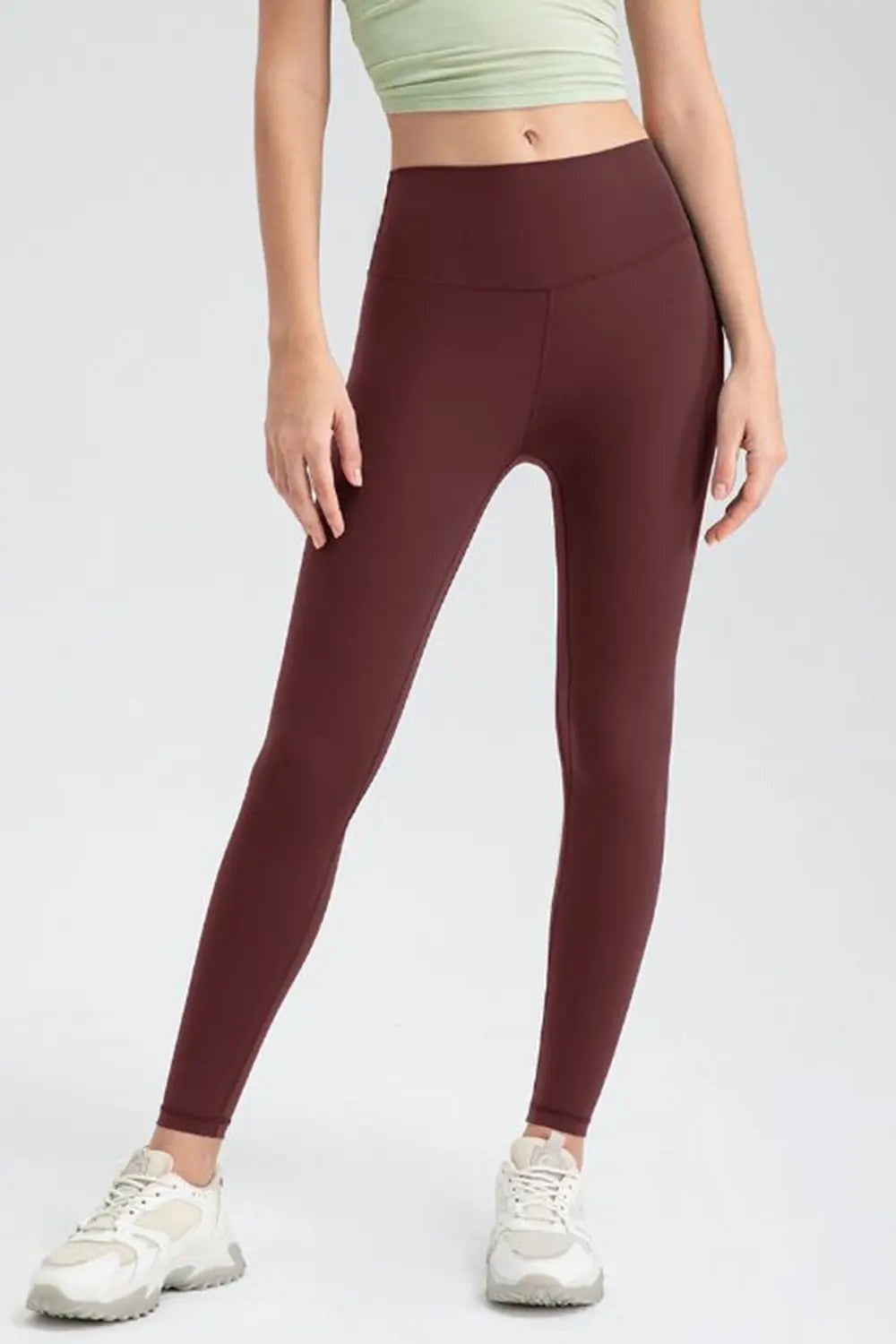 Model wearing burgundy wide-waistband slim-fit active leggings with a high-rise silhouette, smooth construction, and tapered full-length legs, standing with hands at her sides.