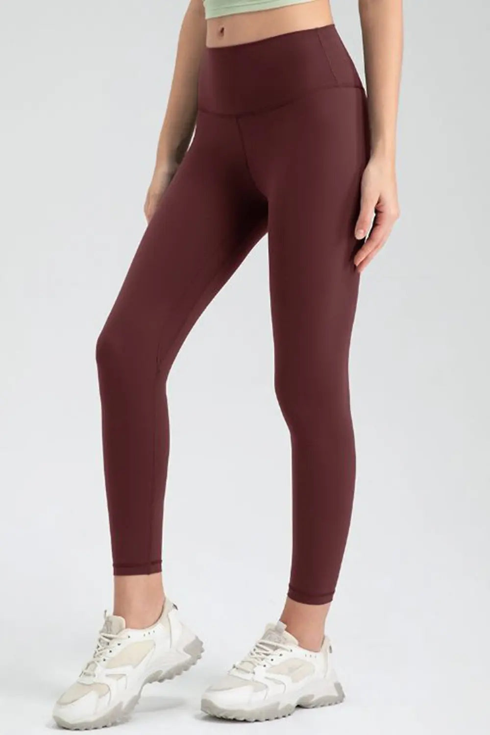 Model poses in burgundy wide-waistband slim-fit active leggings with a high-rise silhouette, stretch construction, ankle-length hems, and visible seam details.