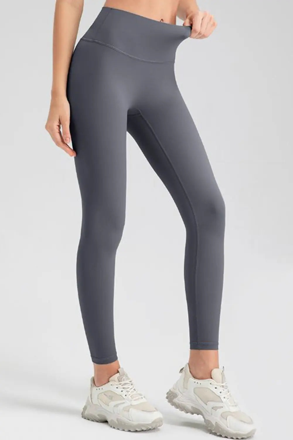 Model wearing dark gray wide-waistband sport leggings with a high-rise silhouette, ankle-length fit, smooth construction, and one hand stretching the waistband.