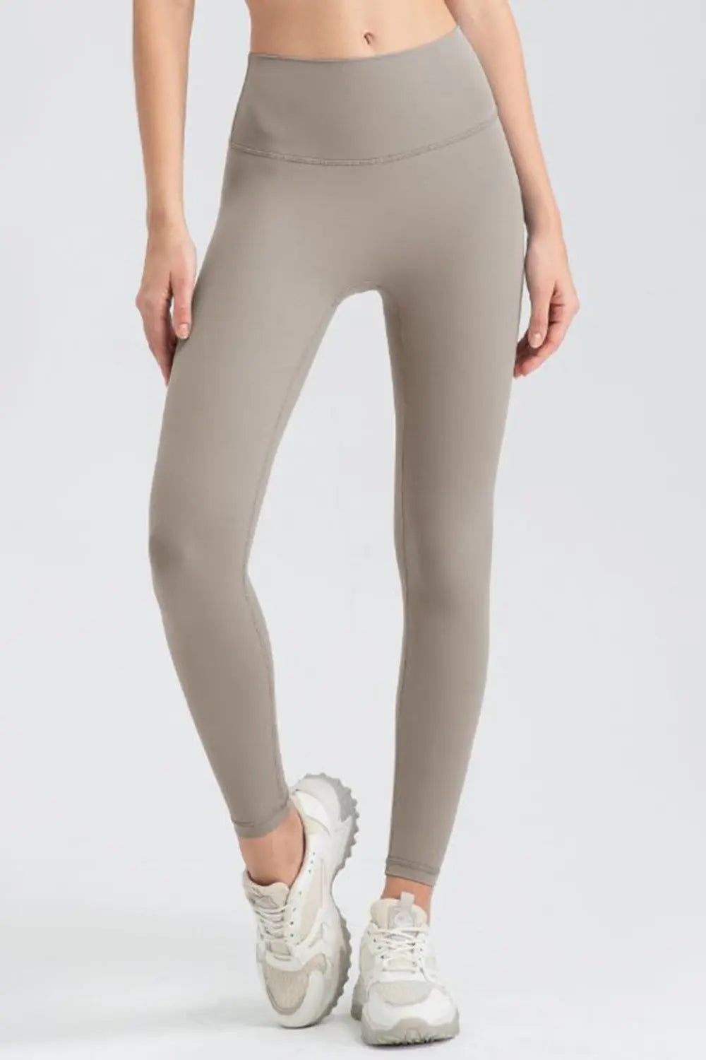 Model wears taupe wide-waistband sport leggings with a high-rise, smooth fitted silhouette and ankle-length legs, standing with one foot crossed forward.