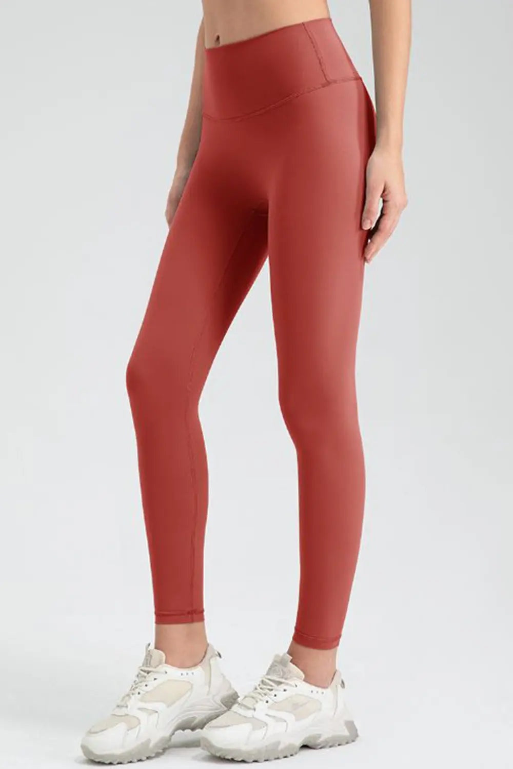 Model stands in coral-red sport leggings with a wide waistband, fitted full-length silhouette, and visible seam construction.
