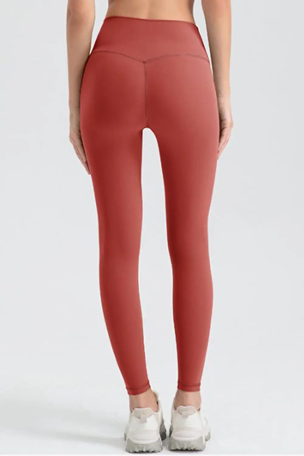 A model stands facing away in rust-red wide waistband sport leggings with a fitted, full-length silhouette and visible center-back seam.