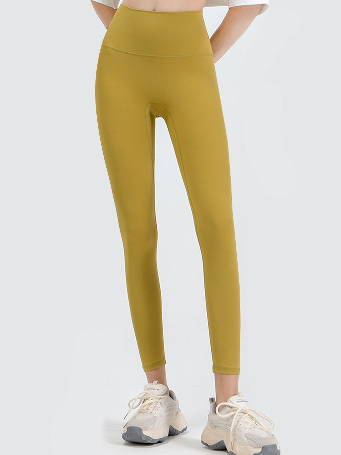 Model stands with hands behind her back wearing mustard yellow wide waistband sports leggings in an opaque, fitted ankle-length silhouette.