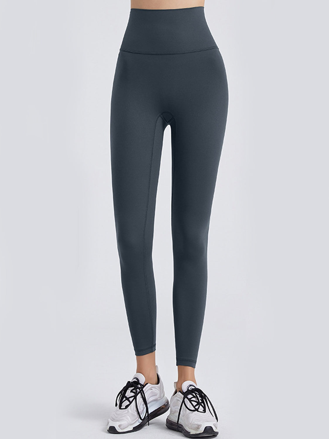 A standing model wears dark charcoal wide waistband sports leggings with a high-rise fit, close tapered silhouette, and visible crotch and inner-leg seams.