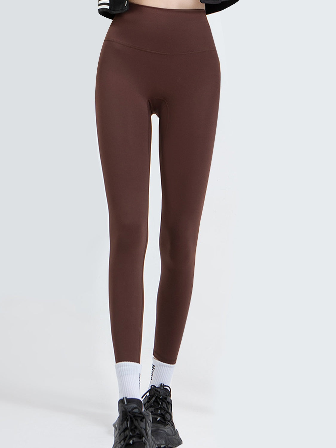 A model wears brown wide waistband sports leggings with a high-rise fit, smooth opaque construction, and a close tapered silhouette while standing with ankles crossed.