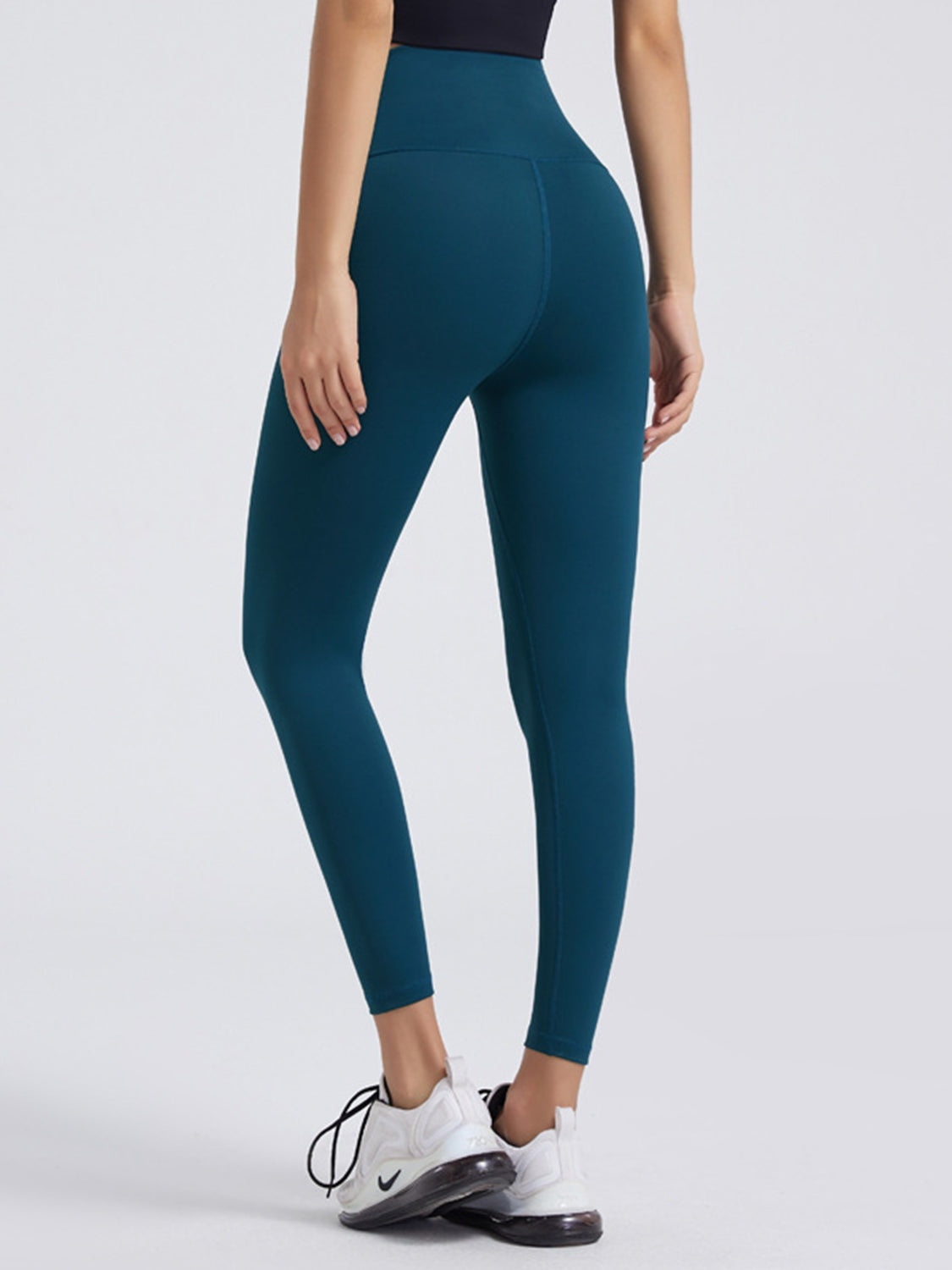 Model shown from behind in dark teal wide waistband sports leggings with a high-rise, fitted ankle-length silhouette and visible curved seam detailing.