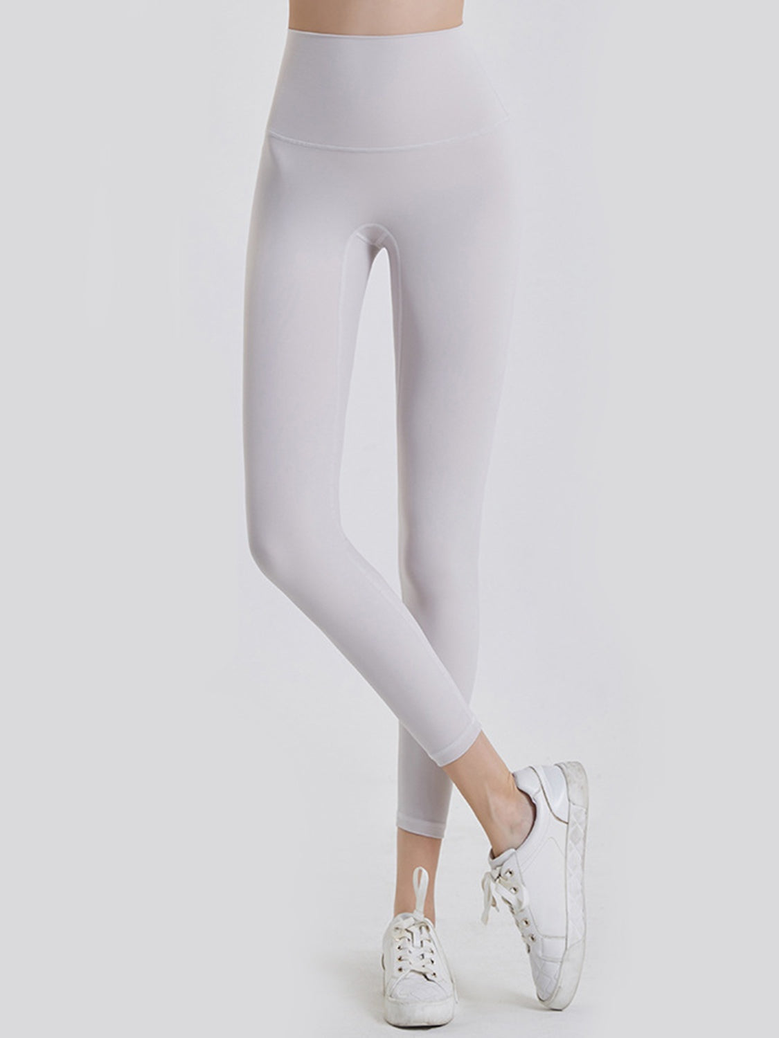 A model poses with one leg crossed over the other in white, opaque sports leggings featuring a wide waistband, fitted ankle-length silhouette, and clean seamed construction.