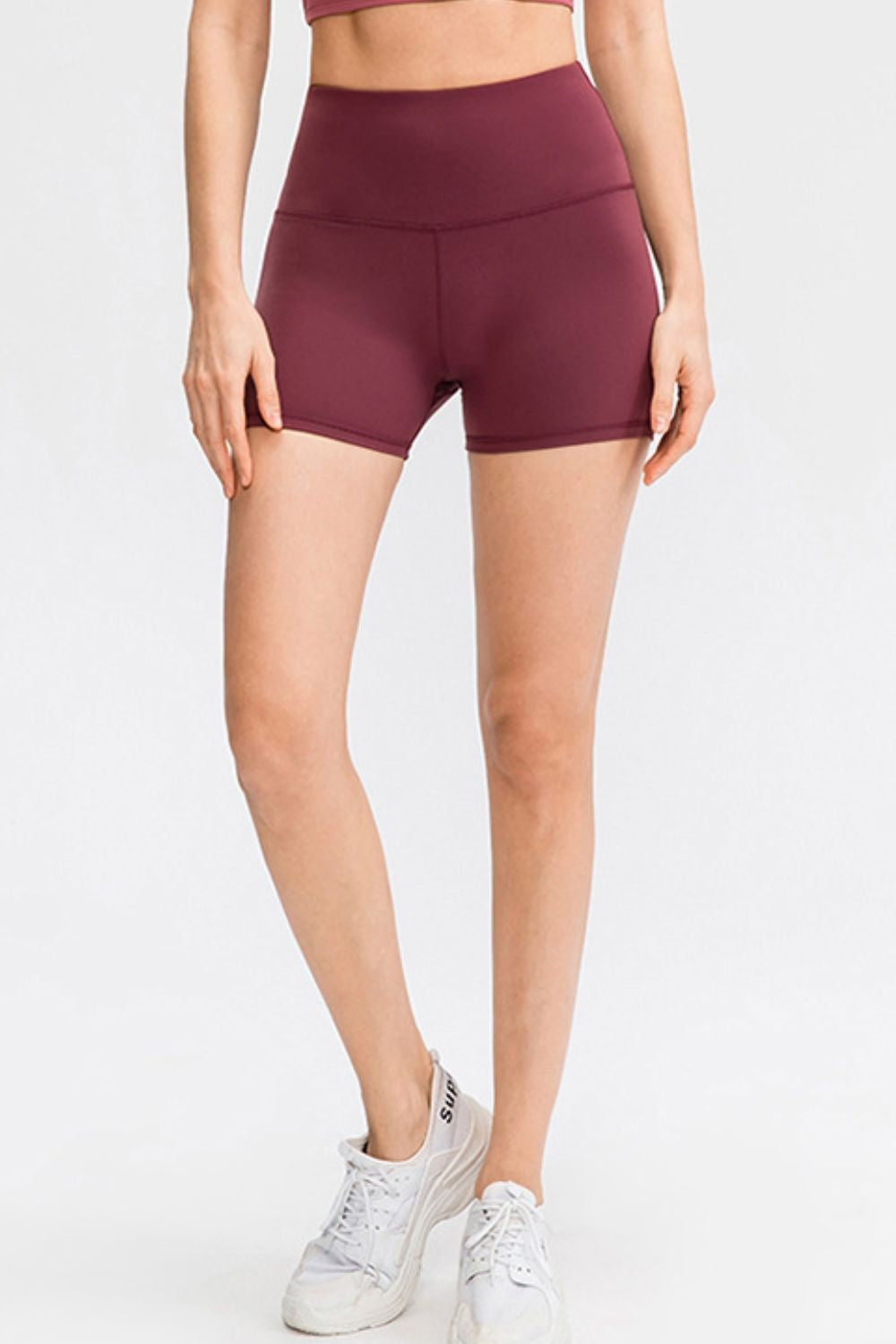 A model wears solid burgundy sports shorts with a wide waistband, fitted mid-thigh silhouette, and visible center seam, standing with ankles crossed and hands at her sides.