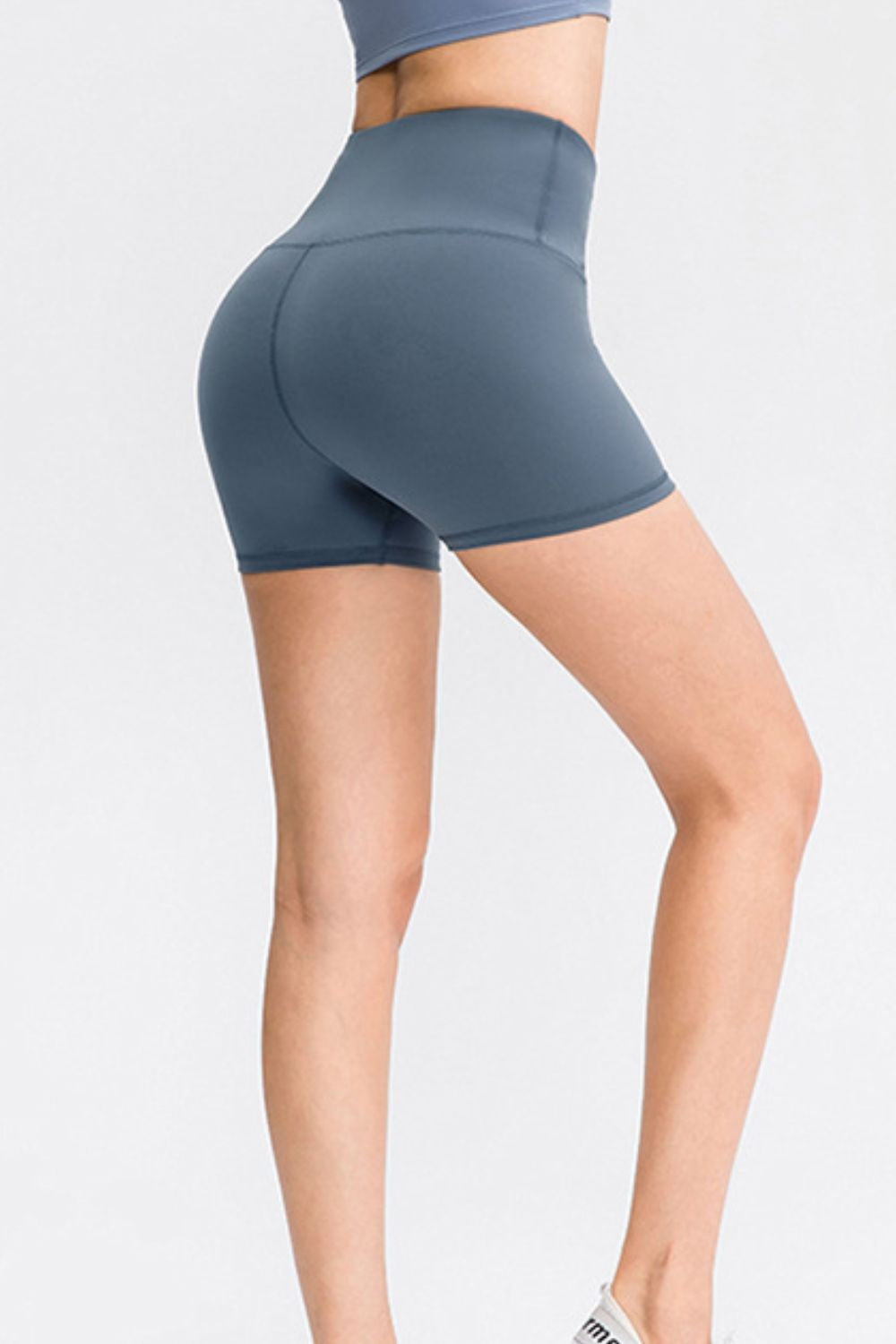 Model wears solid blue-gray wide waistband sports shorts with a high-rise fit, fitted mid-thigh silhouette, and visible seam construction while standing with one leg bent.