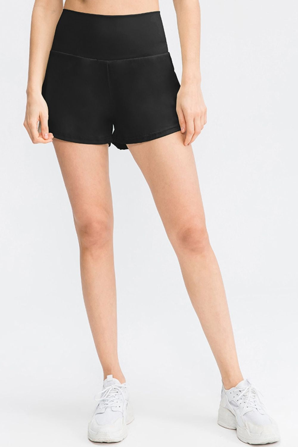 Model stands with legs apart wearing solid black sporty shorts with a wide waistband, moderate stretch, curved hem, and visible side pockets.