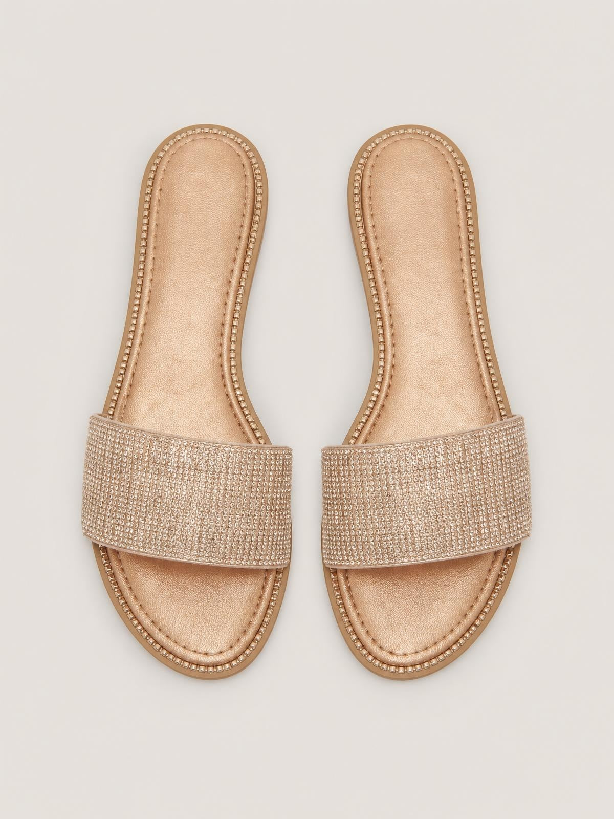 Tan open toe flat sandals feature wide rhinestone-embellished straps, sparkling trim, cushioned-looking insoles, and low flat soles.