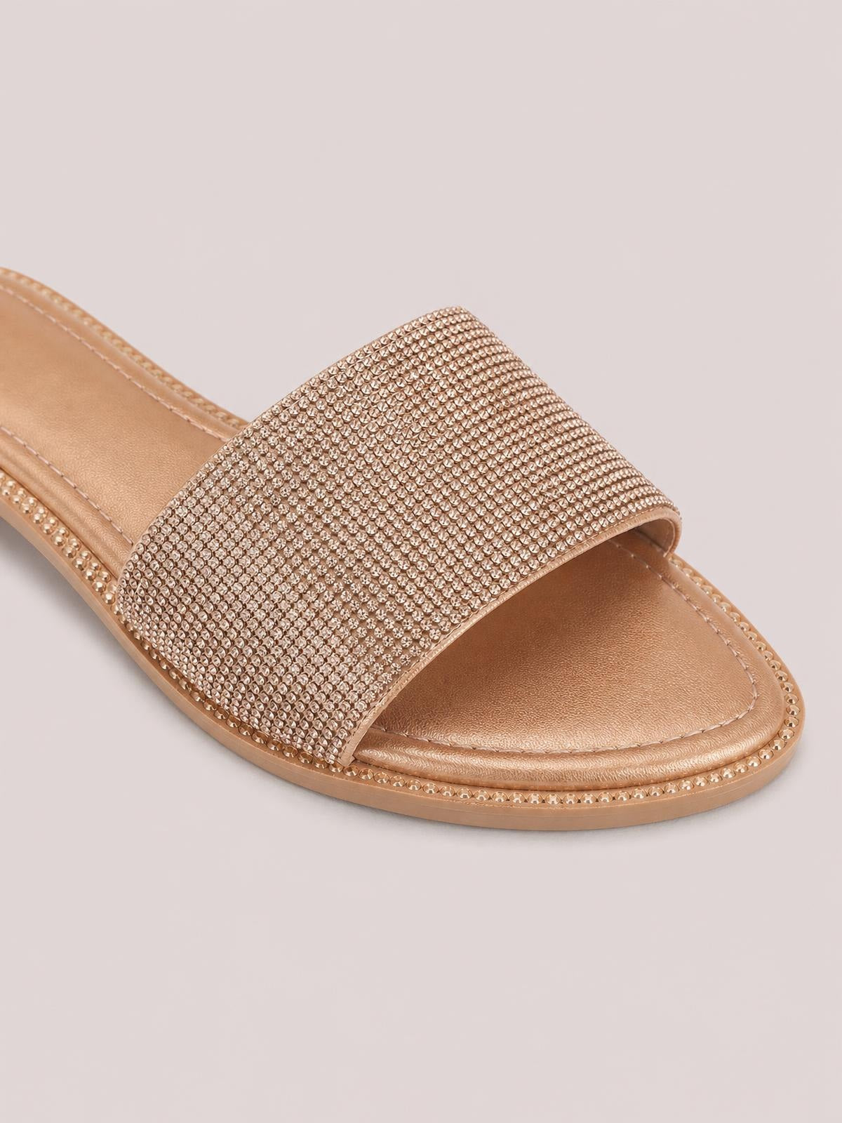 A rose-gold open toe flat sandal features a wide rhinestone-covered band, cushioned footbed, embellished trim, and a low flat sole.