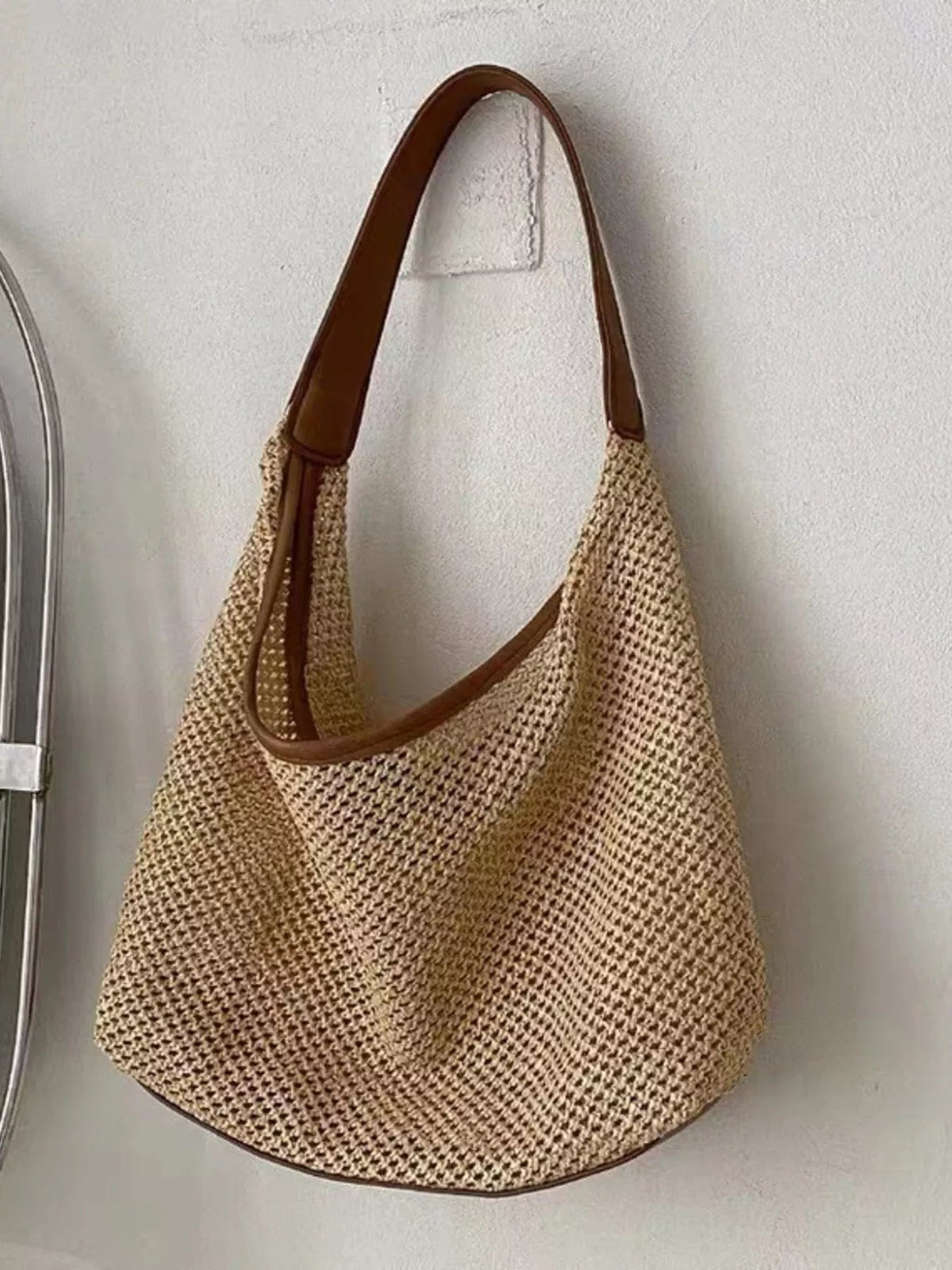 Medium woven straw handbag with a natural beige open-weave body, rounded slouchy silhouette, brown leather trim, and a long shoulder strap.