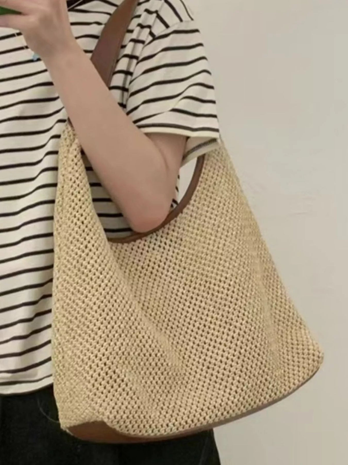 Model holds a medium natural woven straw handbag with a brown leather strap, roomy rounded silhouette, and textured open-knit construction.