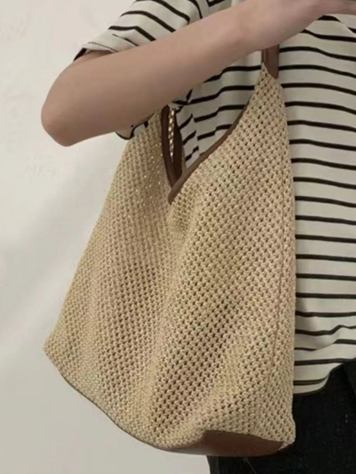 Model holds a medium woven straw handbag with a slouchy silhouette, warm natural color, and brown leather shoulder strap.