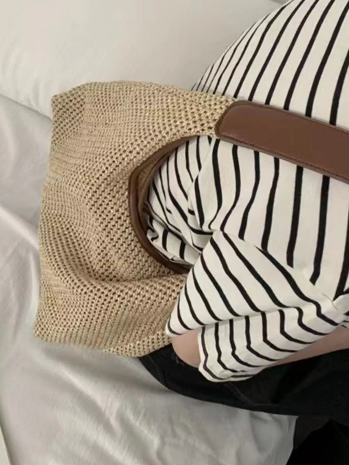 A medium woven straw handbag with a rounded silhouette and brown leather strap rests beside a seated model in a black-and-white striped top and black pants.