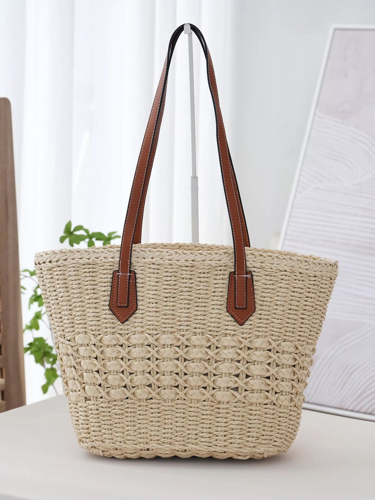 Large woven straw tote bag in a natural beige tone with a structured silhouette, braided texture, and long brown shoulder straps.