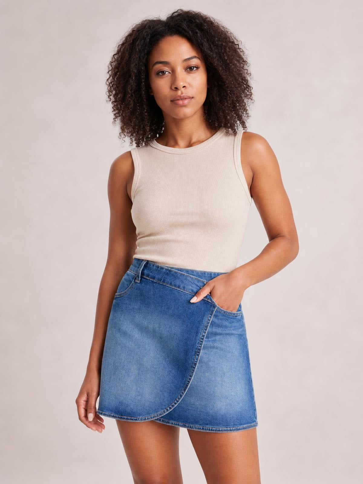 Model poses with one hand in the pocket of a blue denim mini skirt featuring a crossed wrap-style waist, curved overlapping front panel, and straight silhouette.