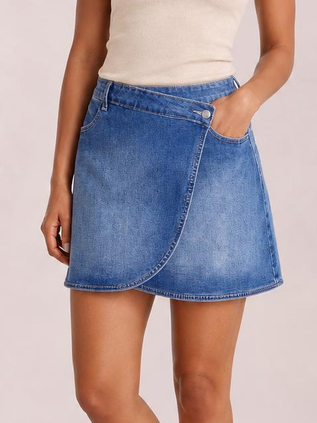 Model wears a blue denim mini skirt with a wrapped crossed waist, curved seam, pocketed design, and relaxed A-line silhouette while posing with one hand in a pocket.