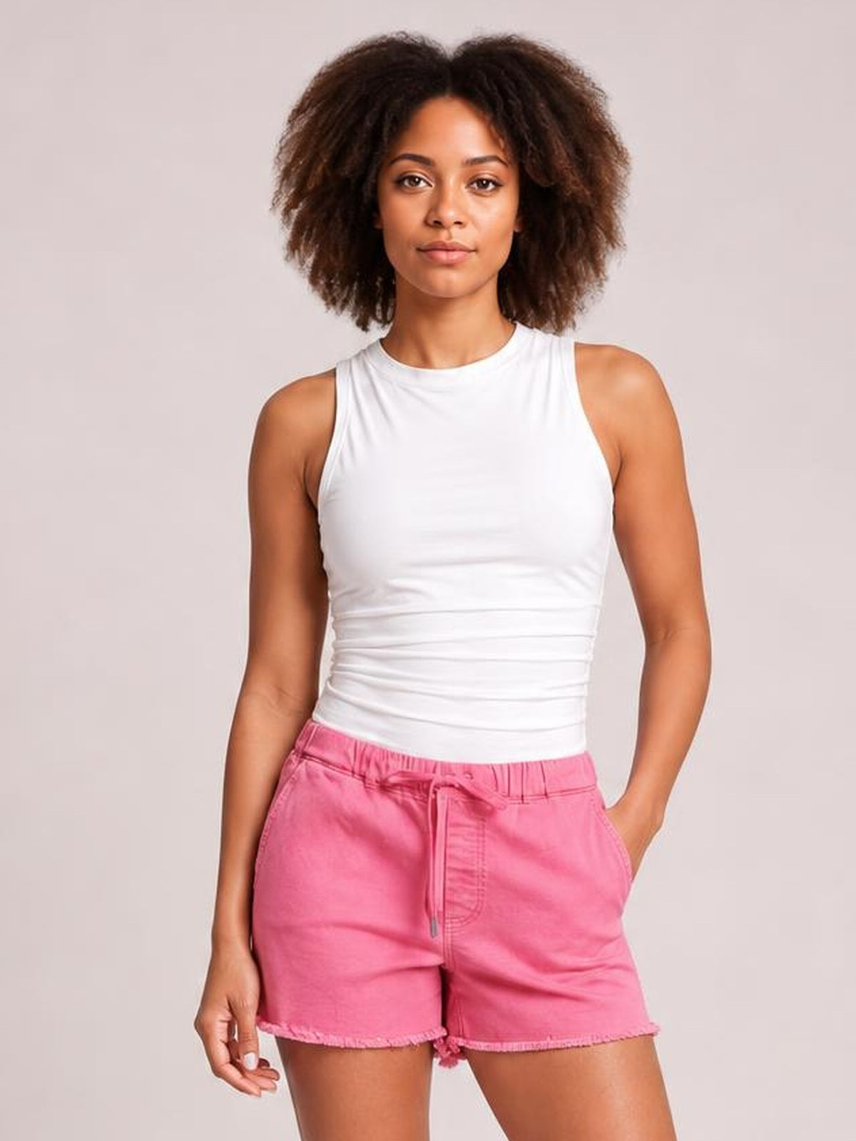 A woman modeling a white sleeveless ribbed tank top and pink denim shorts against a solid light gray studio background.