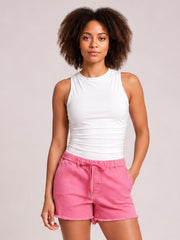 A woman modeling a white sleeveless ribbed tank top and pink denim shorts against a solid light gray studio background.