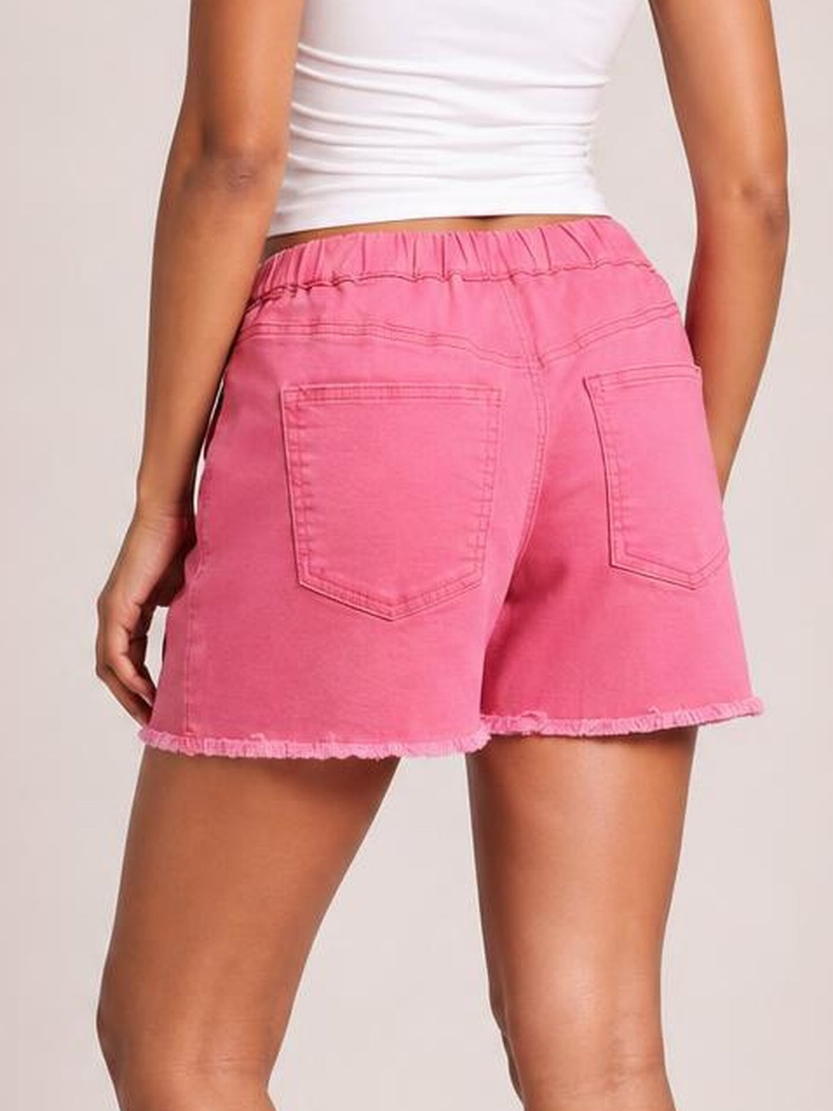 Back view of a model wearing relaxed pink drawstring shorts with an elastic waist, two back pockets, stitched construction, and raw hem edges.