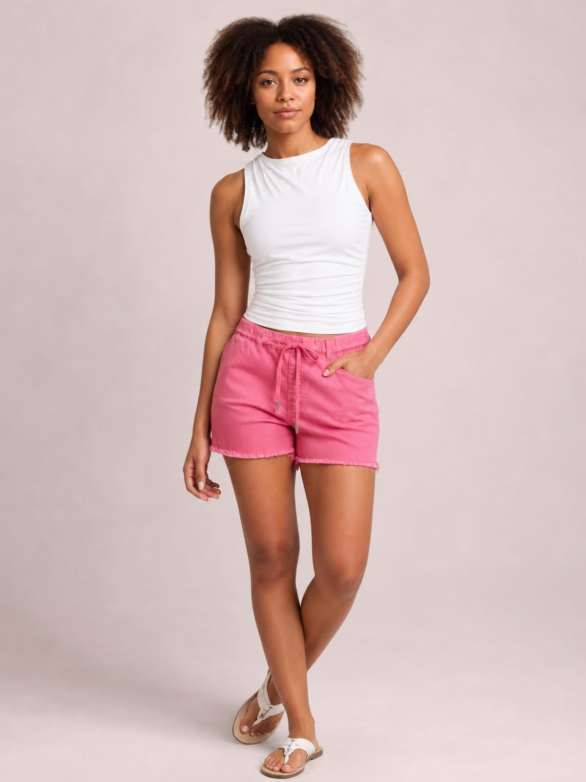 A model poses with one hand in the pocket, wearing relaxed pink drawstring shorts with a comfortable silhouette, front pockets, and raw hem detailing.